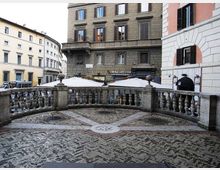 Terrazza urbana pavimentata a motivi geometrici in pietra e mattoni, delimitata da una balaustra in pietra con colonnine e pilastri. Sullo sfondo si vedono palazzi storici con persiane e finestre e alcune coperture bianche di dehors su una strada cittadina.
