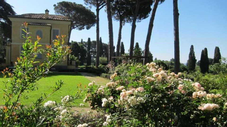 Giardino di una villa con prato curato e aiuole di rose in primo piano, affiancato da un edificio giallo con tetto in tegole. Sullo sfondo si vedono alti pini e cipressi che incorniciano il paesaggio sotto un cielo sereno.