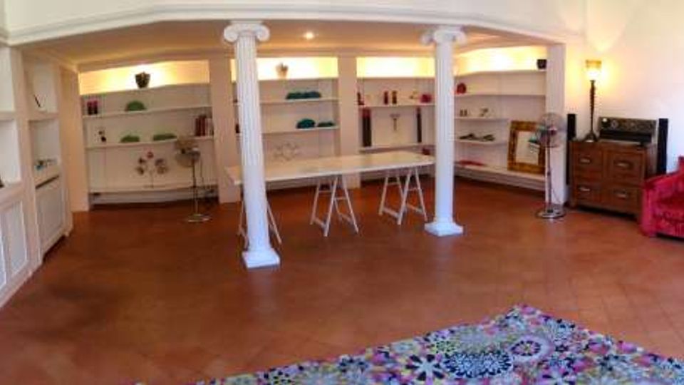 Ampio ambiente interno tipo soggiorno o showroom con pavimento in cotto, scaffalature bianche incassate lungo le pareti e colonne bianche che sostengono un controsoffitto. Illuminazione a parete, alcuni sgabelli e tavolini al centro, con un divano rosso e quadri su una parete laterale.