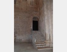 Interno di un edificio storico in pietra con pareti in muratura e una scala di pietra che sale verso un’arcata, conducendo a una porta scura sopraelevata. A destra si vede un grande pilastro in pietra e un pavimento in lastre di pietra.