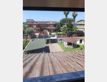 Vista dall’alto su un quartiere residenziale con villette dai tetti in tegole, un vialetto pavimentato con carport e un giardino con gazebo bianco. Sullo sfondo si vedono edifici a due piani, alberi e alte palme sotto un cielo sereno.