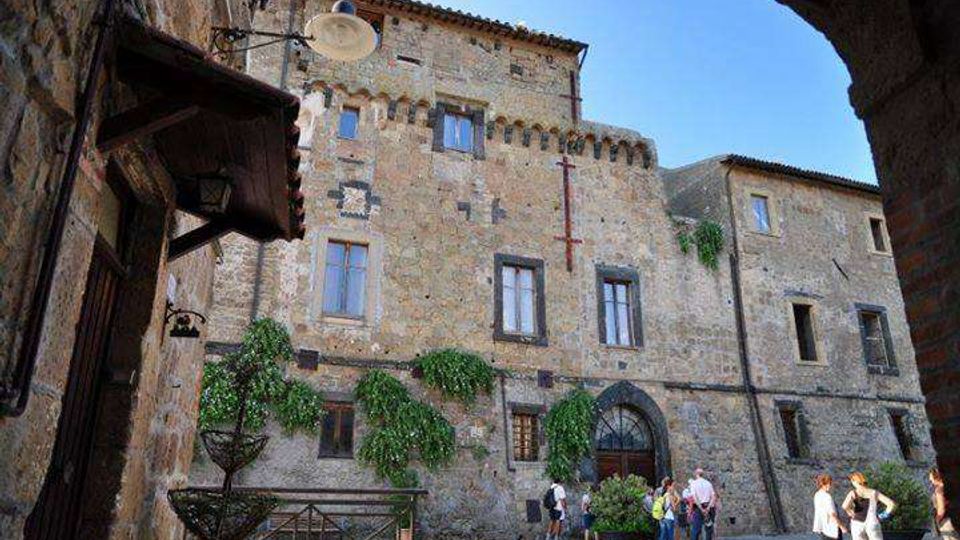 Facciata di un palazzo storico in pietra affacciato su una piccola piazza lastricata, con finestre rettangolari e portale ad arco al centro. La scena è inquadrata da un arco in primo piano e include una lampada a muro e vegetazione rampicante sulle pareti.