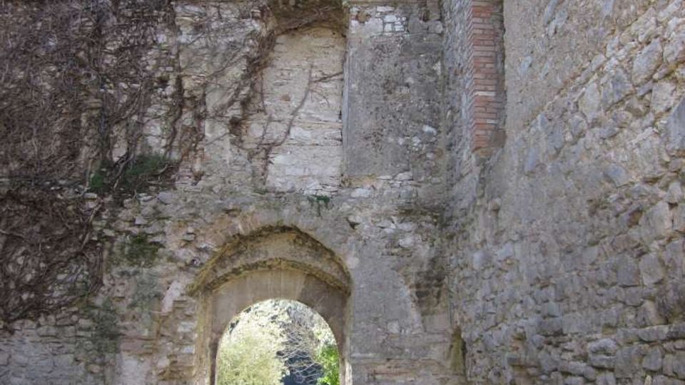 Rovine di una costruzione in pietra con alte mura e un arco d’ingresso, con resti di intonaco e mattoni visibili. Attraverso l’arco si vede un’area erbosa illuminata, circondata da vegetazione e alberi.