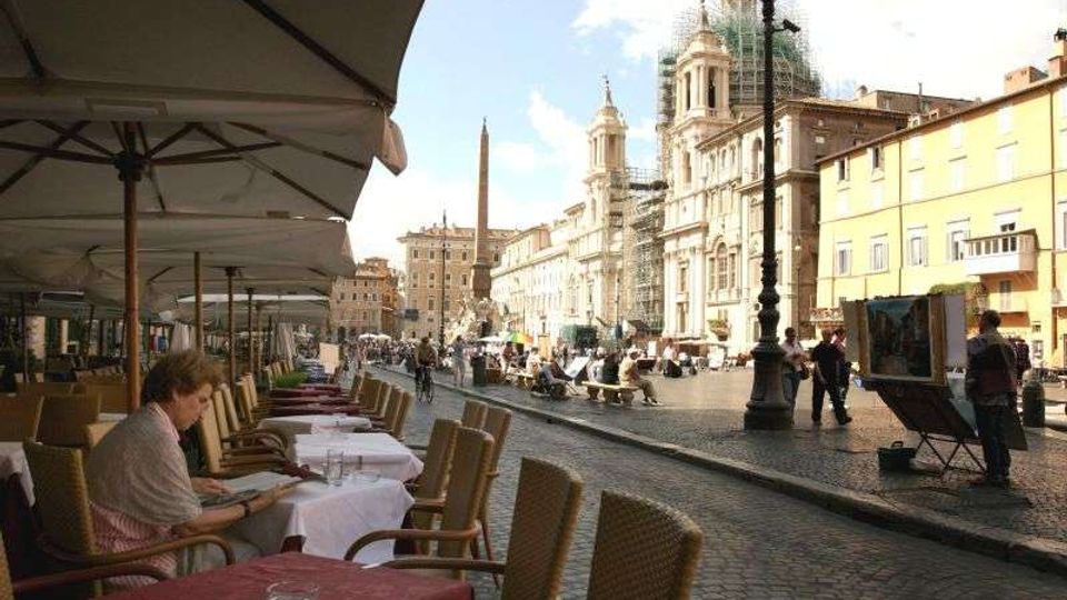 Piazza urbana con pavimentazione in ciottoli, tavolini e sedie di un caffè all’aperto sotto grandi ombrelloni lungo il bordo sinistro. Sullo sfondo si vede una chiesa barocca con cupola (parzialmente coperta da impalcature), un obelisco al centro della piazza e palazzi storici illuminati dal sole.
