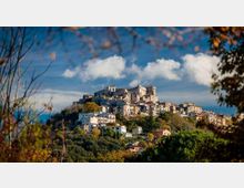 Borgo collinare con case in pietra e tetti chiari disposte a terrazze, dominato da un grande castello-fortezza con torri e mura sulla sommità. In primo piano vegetazione e alberi, con cielo azzurro e nuvole sullo sfondo.