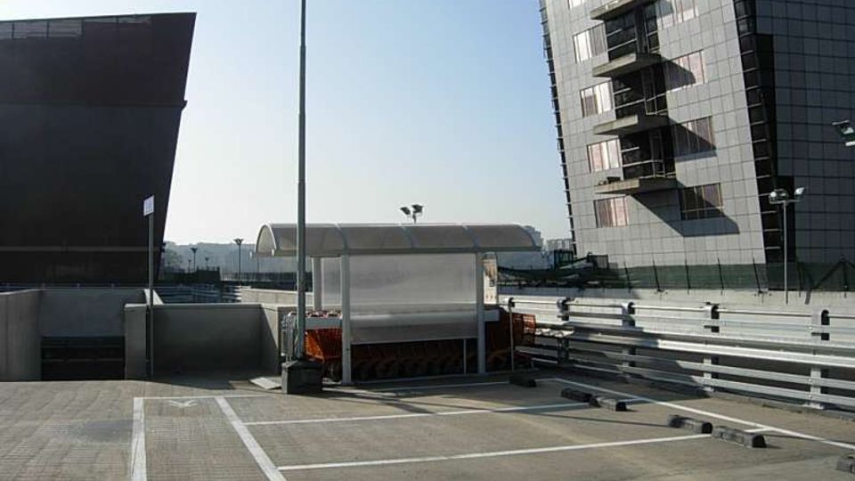 Parcheggio scoperto con stalli delimitati da linee bianche, lampioni alti e una pensilina trasparente al centro. Sullo sfondo si vedono un edificio moderno a più piani con facciata scura e balconi e una lunga struttura arcuata tipo capannone.