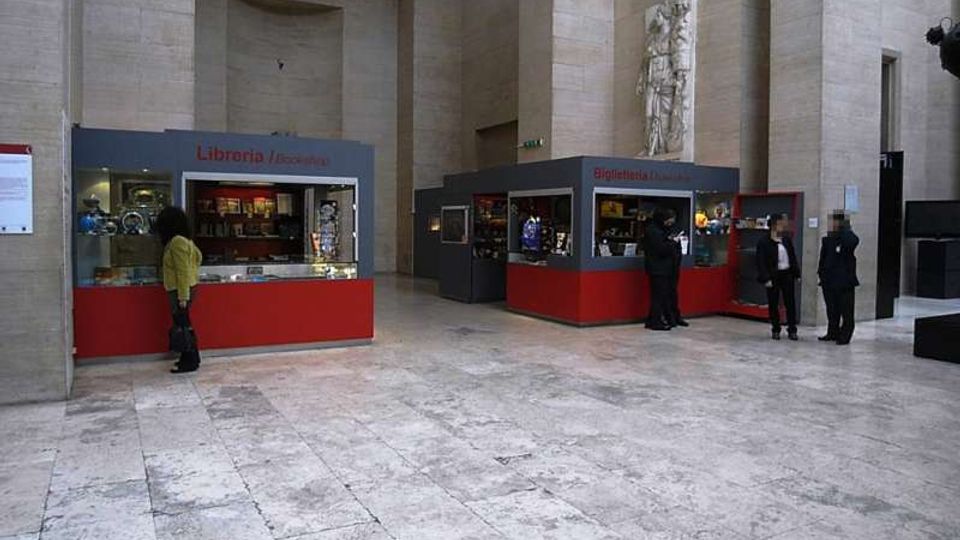 Ampio atrio interno di un edificio in pietra chiara con pavimento in lastre e pareti alte. Nello spazio sono presenti due chioschi con vetrine rosse e grigie per libreria e biglietteria, con una scultura murale in rilievo su una parete.