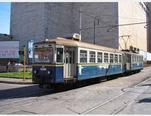 Tram blu e crema su rotaie in una strada urbana, con cavi elettrici aerei e palo della linea. Sullo sfondo si vedono grandi edifici in cemento e un marciapiede con segnaletica e cartelloni.