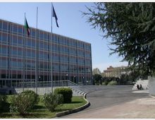 Edificio pubblico moderno con facciata vetrata a griglia e bandiere su pennoni, affacciato su una piazza aperta con percorso curvo asfaltato e gradinate. Intorno ci sono aiuole e alberi, con altri edifici visibili sullo sfondo.