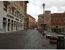 Strada urbana lastricata con palazzi in muratura su entrambi i lati, lampione centrale e marciapiede con panchine e fioriere. A sinistra si vede un edificio con grandi vetrine e l’insegna “Azienda Elettrica Romana”, mentre sulla destra c’è una ringhiera lungo un’area rialzata con altri edifici sullo sfondo.