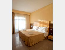Camera da letto d’albergo con letto matrimoniale, testiera in legno e comodino, pavimento piastrellato e pareti color beige. Una grande finestra con tende lascia entrare luce naturale.