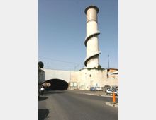 Torre cilindrica in cemento con scala a chiocciola esterna, accanto a un sottopasso stradale sotto un viadotto. L’area circostante è urbana e industriale, con muri in cemento con graffiti, parcheggio di auto e linee ferroviarie sopraelevate.