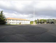Ampio piazzale asfaltato con al centro alti pennoni portabandiera, davanti a un edificio basso e allungato a due piani con tetto in coppi e facciata chiara. Sullo sfondo si vedono alberi e altre costruzioni, sotto un cielo nuvoloso.