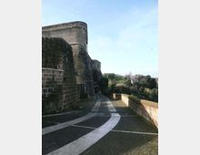 Camminamento in pavé lungo le mura di una fortificazione in pietra, con torre curva e pareti alte ricoperte in parte di vegetazione. Il percorso è affiancato da un parapetto in muratura e si apre verso un panorama di alberi e case sullo sfondo.