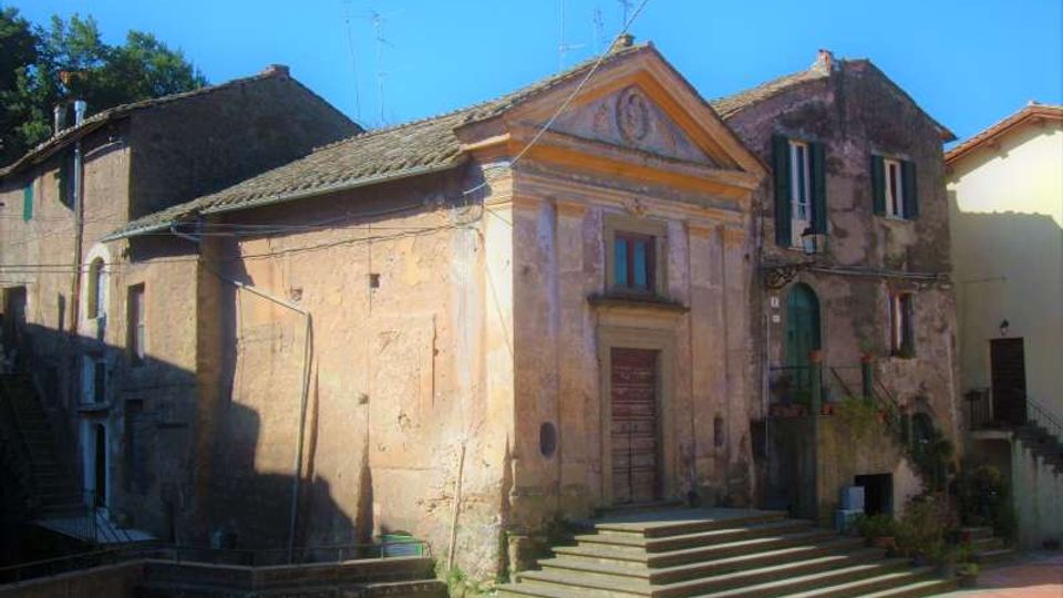 Piccola chiesa in muratura intonacata con facciata a timpano e portale centrale, affiancata da edifici in pietra. Davanti si trova una piazza lastricata con ampia scalinata che conduce all’ingresso, sotto un cielo sereno con cavi aerei visibili.