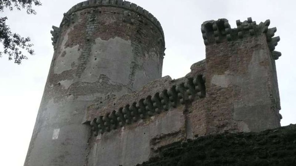 Vista dal basso di una fortificazione in muratura con una torre cilindrica e un mastio rettangolare, entrambi con merlature e beccatelli sporgenti. Le pareti mostrano intonaco consumato e mattoni a vista, sopra una scarpata rocciosa con rami d’albero ai margini del cielo.