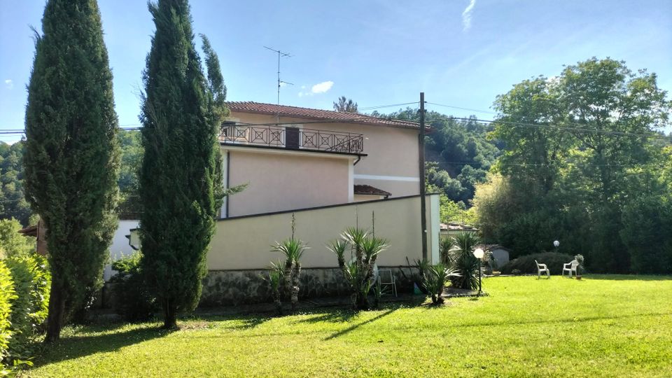Ampio prato verde in un giardino di campagna con cipressi alti, cespugli e piccole palme lungo un muretto. Sullo sfondo si vede una casa a due piani con tetto in tegole e balcone, circondata da alberi e colline boscose.