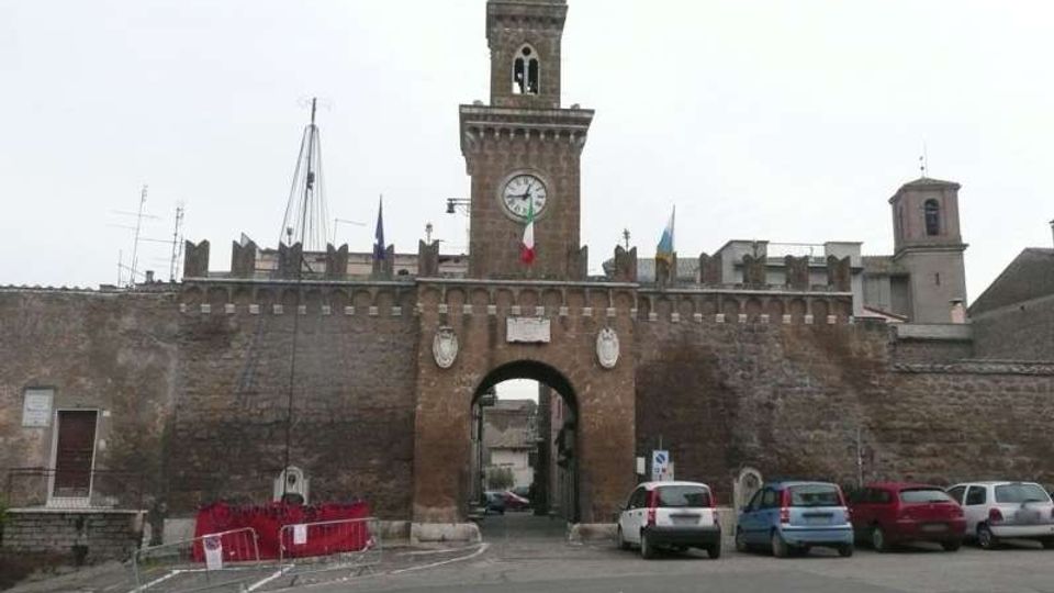 Porta cittadina fortificata in mattoni con grande arco centrale e torre merlata con orologio sulla facciata. Davanti c’è una piccola area stradale con auto parcheggiate e transenne, sotto un cielo coperto.
