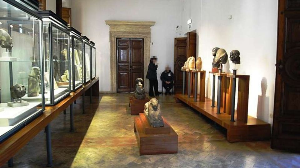 Interno di una sala museale con pareti bianche, pavimento in pietra e faretti a soffitto, con una grande porta in legno incorniciata da un portale in pietra sul fondo. Lungo la parete sinistra ci sono teche in vetro su supporti, mentre a destra e al centro sono esposte sculture e busti su piedistalli e basamenti in legno.