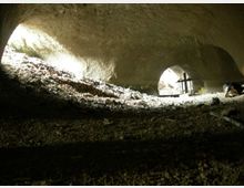 Interno di una grotta naturale con soffitto roccioso basso e irregolare, illuminata da aperture laterali che lasciano entrare fasci di luce. Il pavimento è ricoperto di terra e detriti, con un piccolo tavolo e oggetti visibili in lontananza vicino all’uscita.