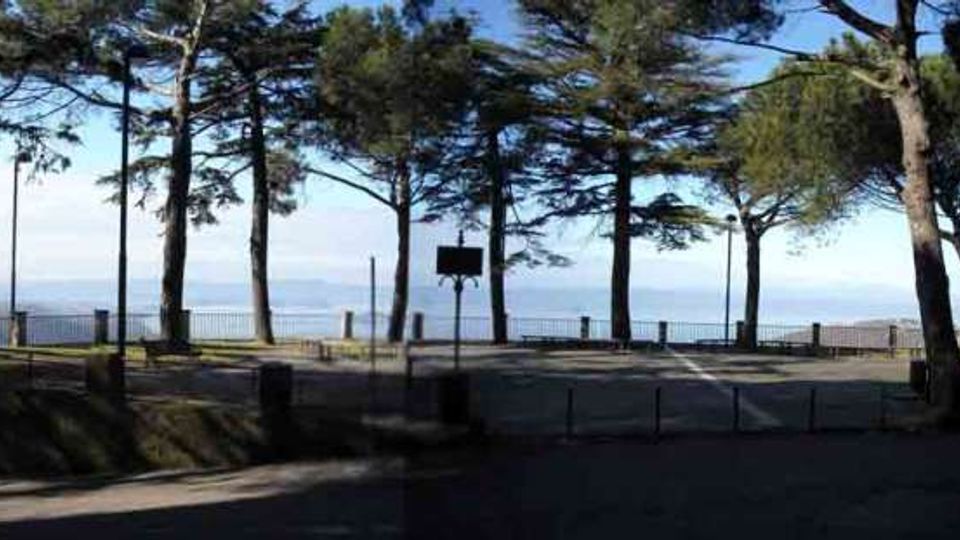 Spiazzo panoramico alberato con pini alti e ringhiera lungo il bordo, affacciato sul mare all’orizzonte. Area pavimentata con cartello informativo e strada in primo piano.