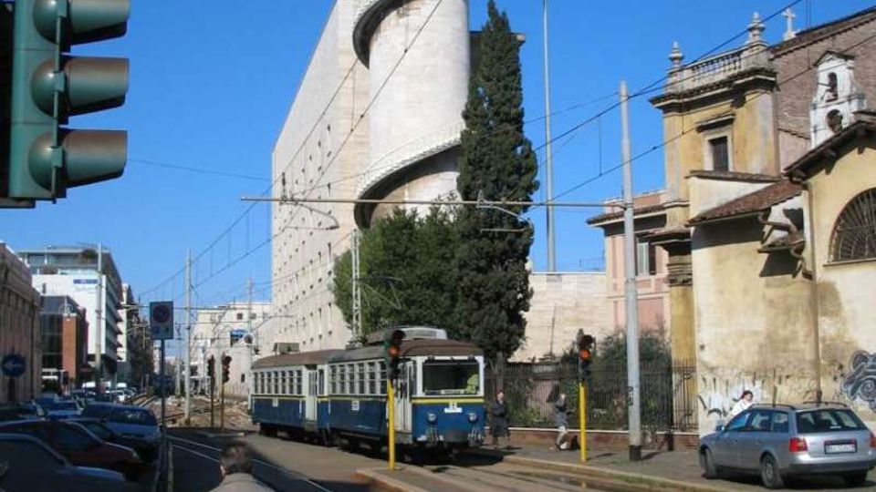 Strada urbana con binari del tram e linea aerea, affiancata da edifici e auto parcheggiate. Sullo sfondo si vede una torre cilindrica in cemento con una rampa elicoidale esterna e un tram che percorre la carreggiata.