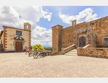 Piazza lastricata in pietra con una piccola chiesa con campanile a vela e, a destra, un grande edificio fortificato in muratura con torre e portale ad arco raggiunto da una rampa con parapetti. Sullo sfondo si vedono alberi e colline sotto un cielo azzurro con nuvole.