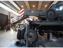 Interno di un’officina o capannone industriale con pavimento in cemento e tetto in metallo illuminato da luci a soffitto. In primo piano si vede un grande veicolo gru in manutenzione, con braccio e stabilizzatori con bande rosse e bianche, ruote e componenti meccanici sparsi a terra.