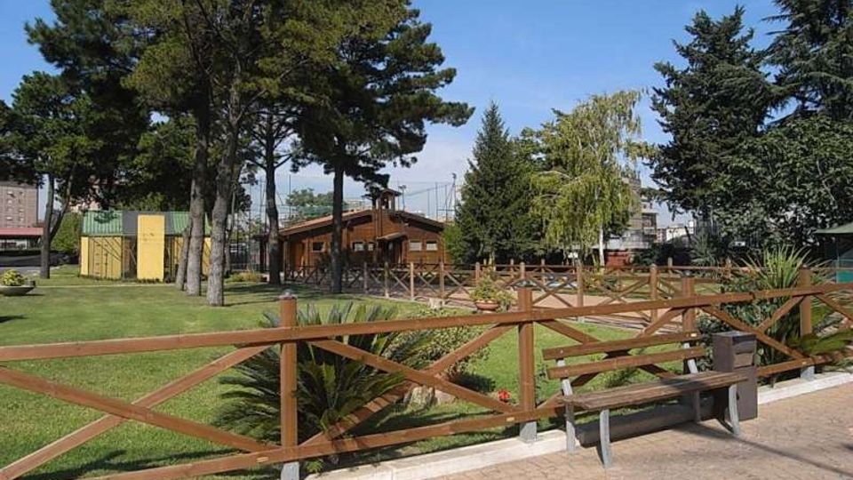 Parco urbano con ampio prato e alberi alti, delimitato da una staccionata in legno lungo un vialetto pavimentato con panchina. Sullo sfondo si vedono una piccola struttura in legno e un’area recintata con edifici vicini.