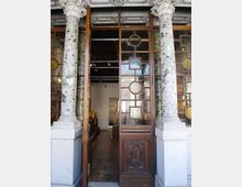 Ingresso di un edificio storico con due colonne in marmo maculato e capitelli decorati, che incorniciano una grande porta in legno con pannelli di vetro geometrici. Attraverso la porta aperta si intravede un interno tipo galleria o museo con pavimento scuro, illuminazione a soffitto e sculture esposte.