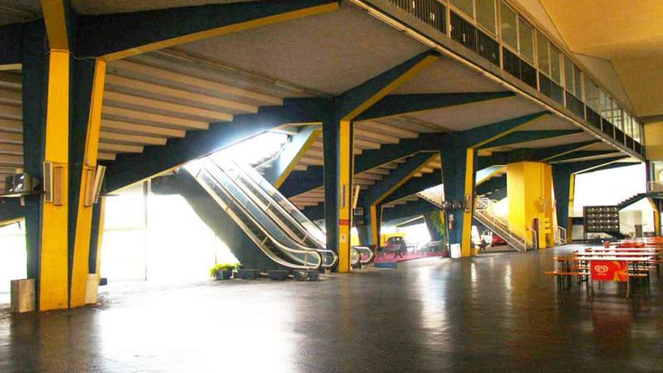 Ampio spazio interno di una struttura moderna con grandi pilastri gialli e travi a vista, pavimentazione scura e soffitto alto. Due scale mobili e una passerella sopraelevata con ringhiera conducono al livello superiore, con luce intensa che entra da aperture laterali.