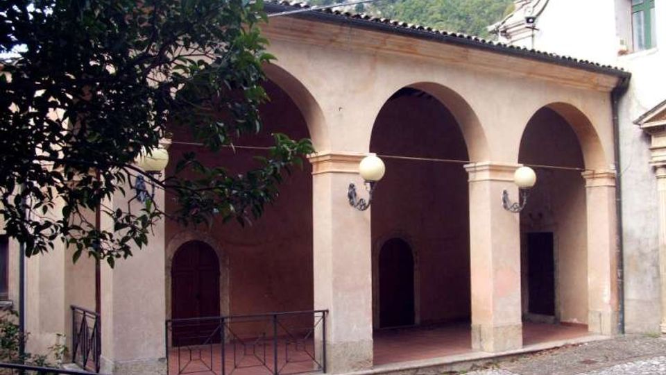 Portico di un edificio in muratura intonacata con tre grandi arcate e colonne squadrate, pavimento in cotto e lampioni a sfera fissati alle pareti. In primo piano si vede una piccola area lastricata con ringhiera metallica e un albero, con colline verdi sullo sfondo.