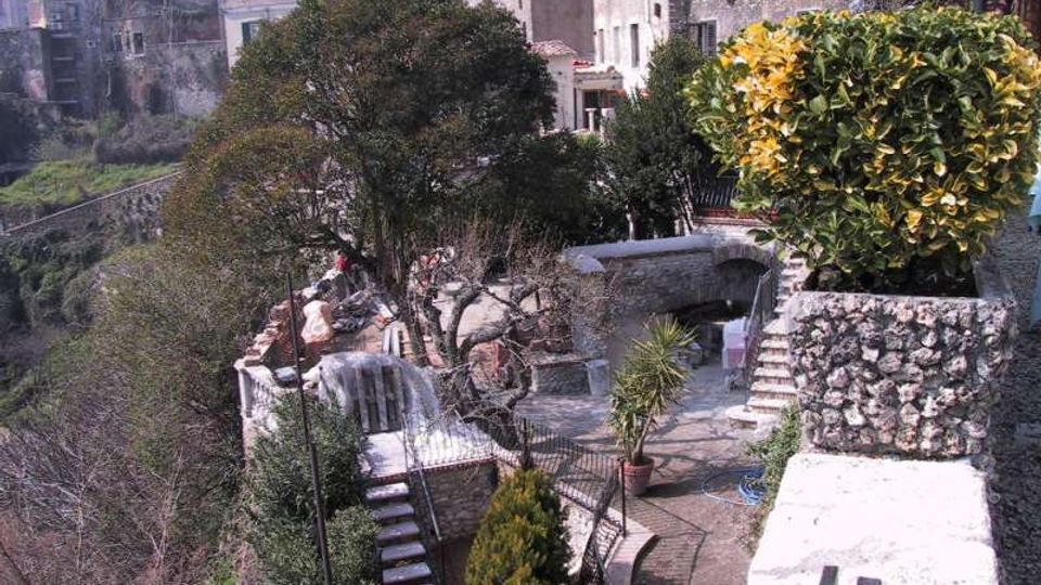 Terrazza panoramica in pietra su un pendio, con muretti, scale e ringhiere che collegano piccoli terrazzamenti con alberi e piante in vaso. Sullo sfondo si vedono case addossate tra loro lungo il bordo del borgo e la valle sottostante.