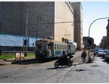 Strada urbana con binari del tram al centro e cavi aerei, con un tram blu in transito accanto a un grande edificio moderno in cemento. Sul lato destro si vedono auto in corsia e lampioni e semafori lungo il marciapiede.