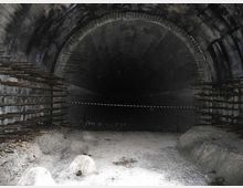Interno di una galleria o tunnel in cemento con volta semicircolare, pareti annerite e barre metalliche sporgenti sui lati. Il pavimento è coperto di detriti e l’apertura verso il fondo appare buia e profonda.