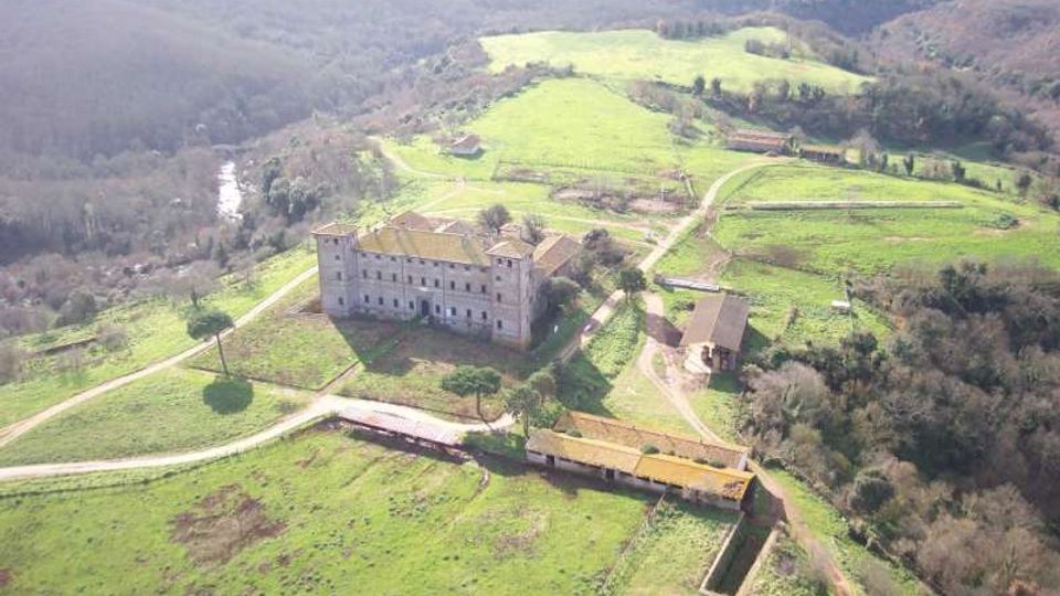 Veduta aerea di un grande edificio storico in pietra con torre, circondato da prati verdi e strade sterrate in una campagna collinare. Intorno si vedono campi, piccoli edifici rurali e un’area boschiva ai margini.