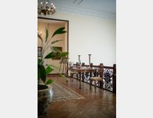 Interno di un salone in stile classico con pavimento in parquet, soffitto decorato in stucco e lampadario; lungo una ringhiera in ferro battuto ci sono un tavolo con ceramiche e candelieri e sedie imbottite. Sullo sfondo si vede un passaggio verso un’altra stanza con quadri e cavalletto, con una pianta in primo piano sfocata.