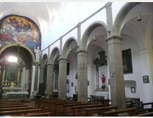 Interno di una chiesa con navata centrale, file di panche in legno e arcate in pietra su colonne che delimitano le navate laterali. Sul fondo si vede l’altare con un grande affresco nell’abside e alcune statue e quadri lungo le pareti bianche.