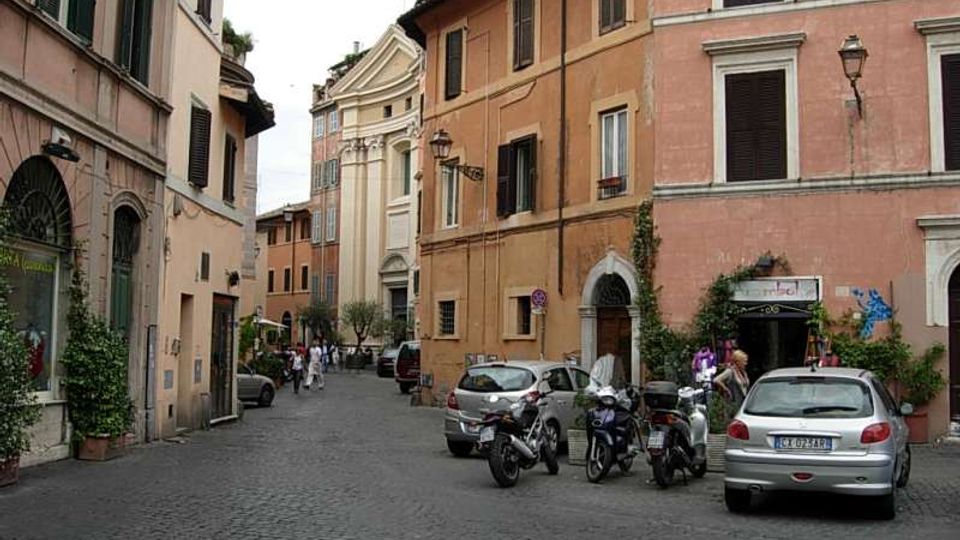 Strada acciottolata in un centro storico con edifici alti dai toni ocra e rosa, finestre con persiane e portali ad arco. Lungo la via sono parcheggiati auto e scooter, e sullo sfondo si vede una facciata chiara in stile classico tra le case.