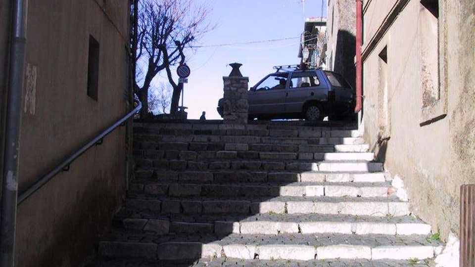 Scalinata in pietra e selciato che sale tra due edifici stretti, con ombre marcate sui gradini. In cima si vedono un lampione, un piccolo muretto in pietra e una strada con un’auto parcheggiata, sotto un cielo sereno.