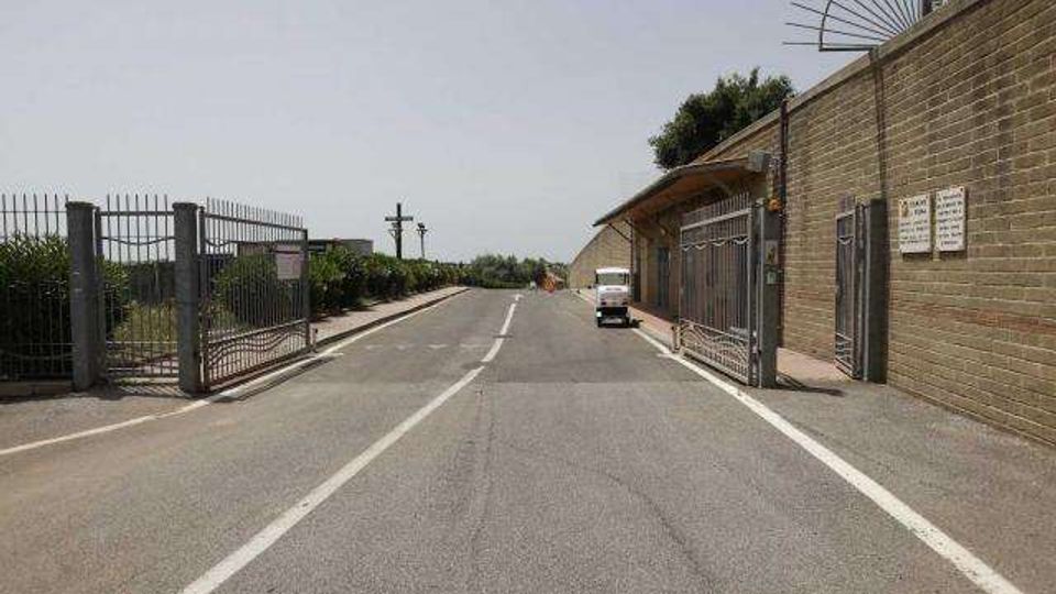 Strada asfaltata a due corsie con linea centrale tratteggiata, affiancata da recinzioni metalliche e da un lungo edificio industriale in mattoni con cancelli e cartelli. Lungo il lato destro è presente un piccolo veicolo parcheggiato e, sullo sfondo, si vedono vegetazione e pali della luce.