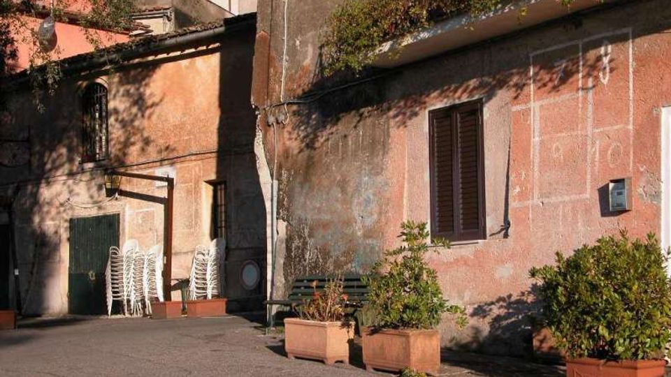 Cortile esterno con pavimentazione in cemento affacciato su edifici intonacati in rosa e grigio, con finestre con persiane, porta verde e muri segnati dal tempo. In primo piano ci sono grandi fioriere con arbusti e, vicino all’ingresso, una pila di sedie bianche impilate.