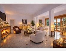 Ampio salone interno con camino acceso in pietra, pavimento in marmo e area lounge con poltrone e divani chiari attorno a un tavolino rotondo. Grandi finestre con infissi in legno illuminano lo spazio, arredato con lampade da tavolo, composizioni floreali e tavoli da buffet con tovaglie drappeggiate.