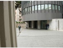 Piazzale urbano pavimentato davanti a un edificio moderno con facciata curva in pannelli metallici e ampie finestre, rialzato su pilastri con ingresso vetrato. Sullo sfondo si vedono un muro in pietra chiara, alberi e palazzi storici adiacenti.