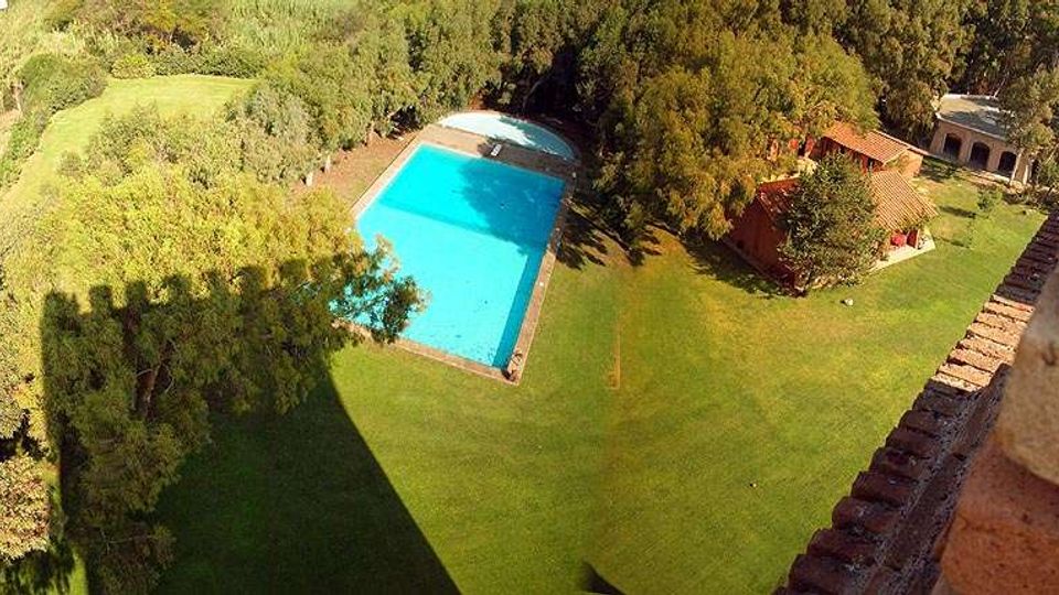 Vista dall’alto di un ampio giardino con grande prato verde e una piscina rettangolare azzurra circondata da alberi. Sul lato destro si vedono edifici con tetto in tegole e portico, e in primo piano un bordo di muratura in mattoni.