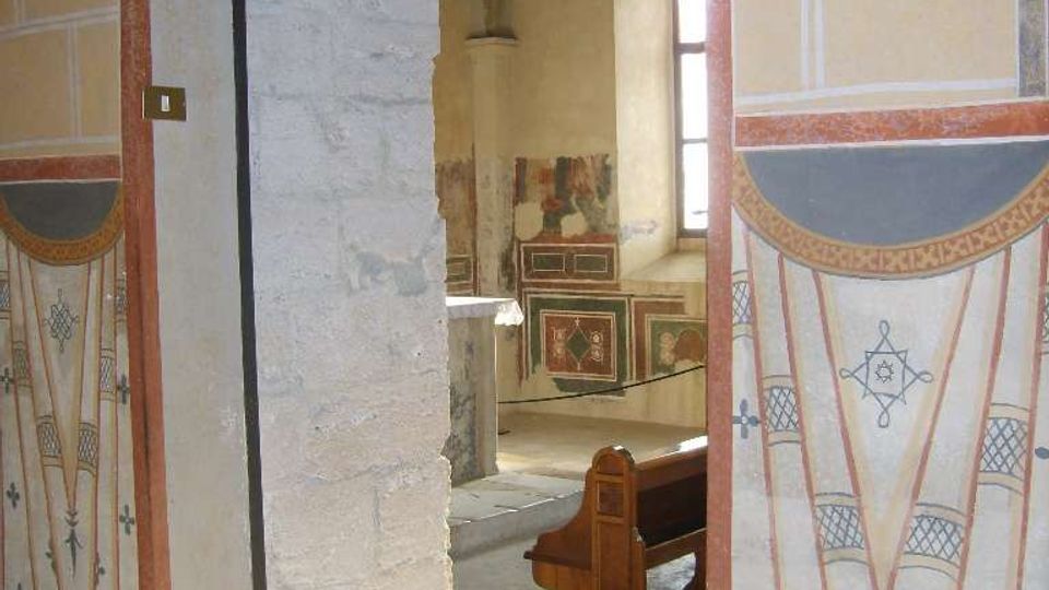 Passaggio ad arco che conduce a un interno di chiesa o cappella, con pareti affrescate e decorazioni geometriche. Si intravedono una finestra ad arco, un banco in legno e pavimento in pietra, con cornici murarie dipinte attorno all’apertura.