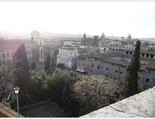 Vista panoramica su un centro storico con edifici in muratura e tetti in tegole, interrotta da cupole e campanili sullo sfondo. In primo piano si vedono alberi alti (cipressi) e una terrazza in pietra che affaccia su un’area verde.