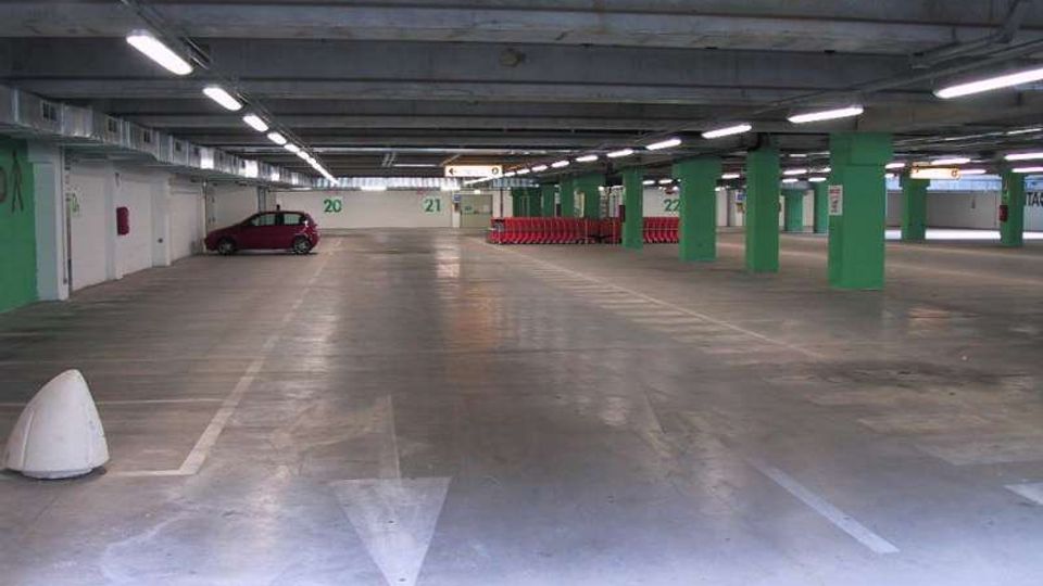 Parcheggio sotterraneo con ampie corsie in cemento, illuminazione a neon e soffitto con tubazioni a vista. Pilastri verdi e segnaletica con numeri dei posti delimitano le aree di sosta.