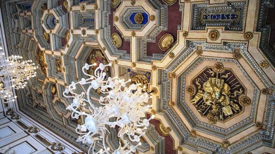 Soffitto interno riccamente decorato con cassettoni ottagonali e riquadri geometrici in rilievo, con dettagli dorati e inserti colorati. Lampadari in vetro sospesi illuminano l’ambiente, evidenziando le cornici e gli elementi ornamentali del soffitto.