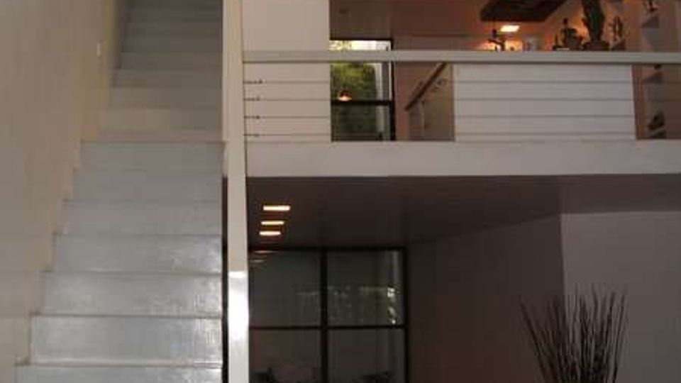 Interno di una casa moderna a doppia altezza con scala bianca laterale che conduce a un soppalco con ringhiera orizzontale. Al piano terra si vede una zona giorno con divano, grande finestra vetrata e una pianta in vaso, mentre al livello superiore appare una cucina con illuminazione a incasso.
